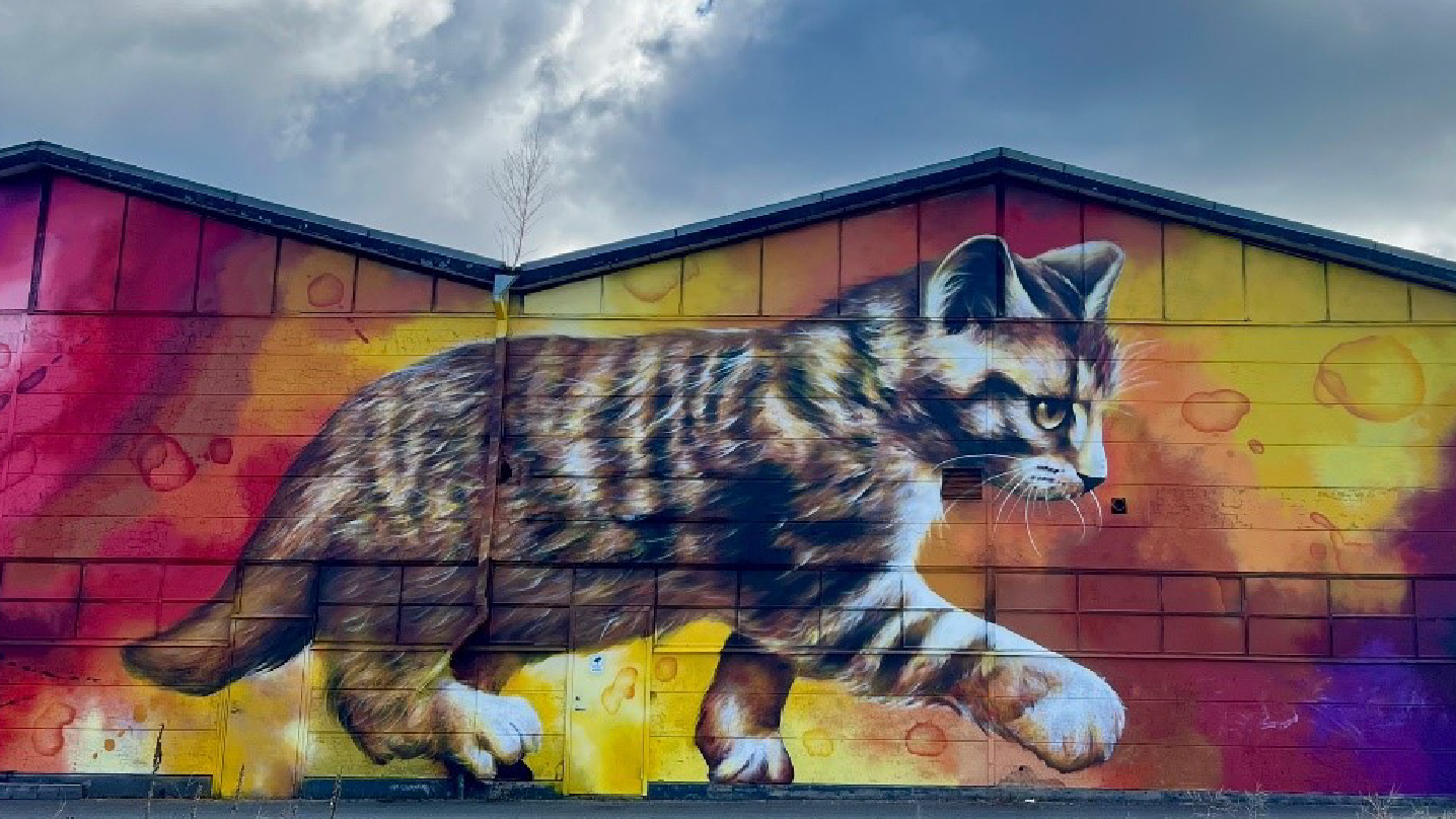 En muralmålning på en gående brun katt. Bakgrunden är i gult, rosa, oranget, rött