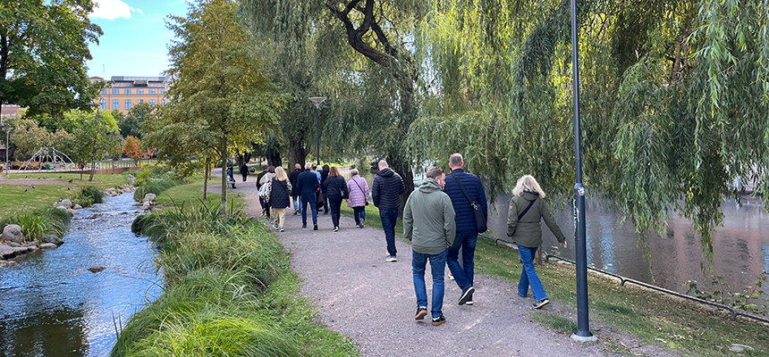 Människor går i en grön lummig park. På höger sida syns en fors och på vänster sida en bäck. Solen lyser på en blå himmel