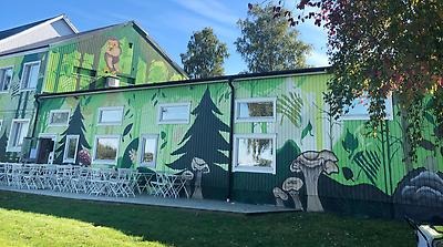 Foto på en del av Kvarteret Ödlans byggnad. Den är fantasifylld graffiti-målad i gröna toner. Blå himmel, träd och gräs syns också. 