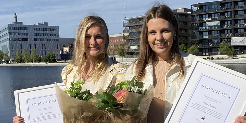 Foto på stipendiater Elly och Anna som håller i blommor samt stipendier. I bakgrunden syns vatten, byggnader och en blå himmel. 