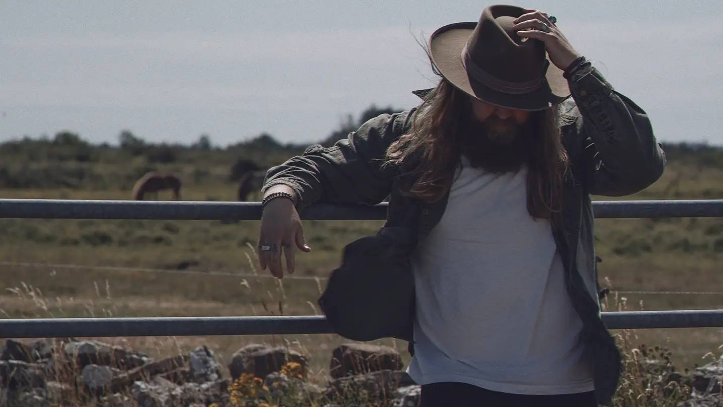 En man med långt hår och cowboyhatt står lutad mot ett staket.