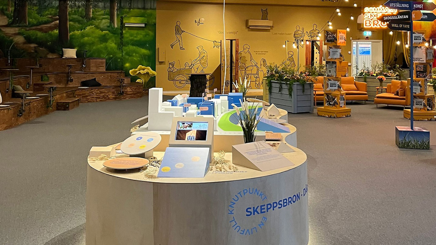 I mitten av ett öppet rum står en stor träform med flera träformer uppepå, formade i olika små byggnader och infoskyltar. Till vänster finns en gradäng formad som ett stort träd, och till höger en orange soffgrupp med massor av blommor runt om och ljusslingor i taket.