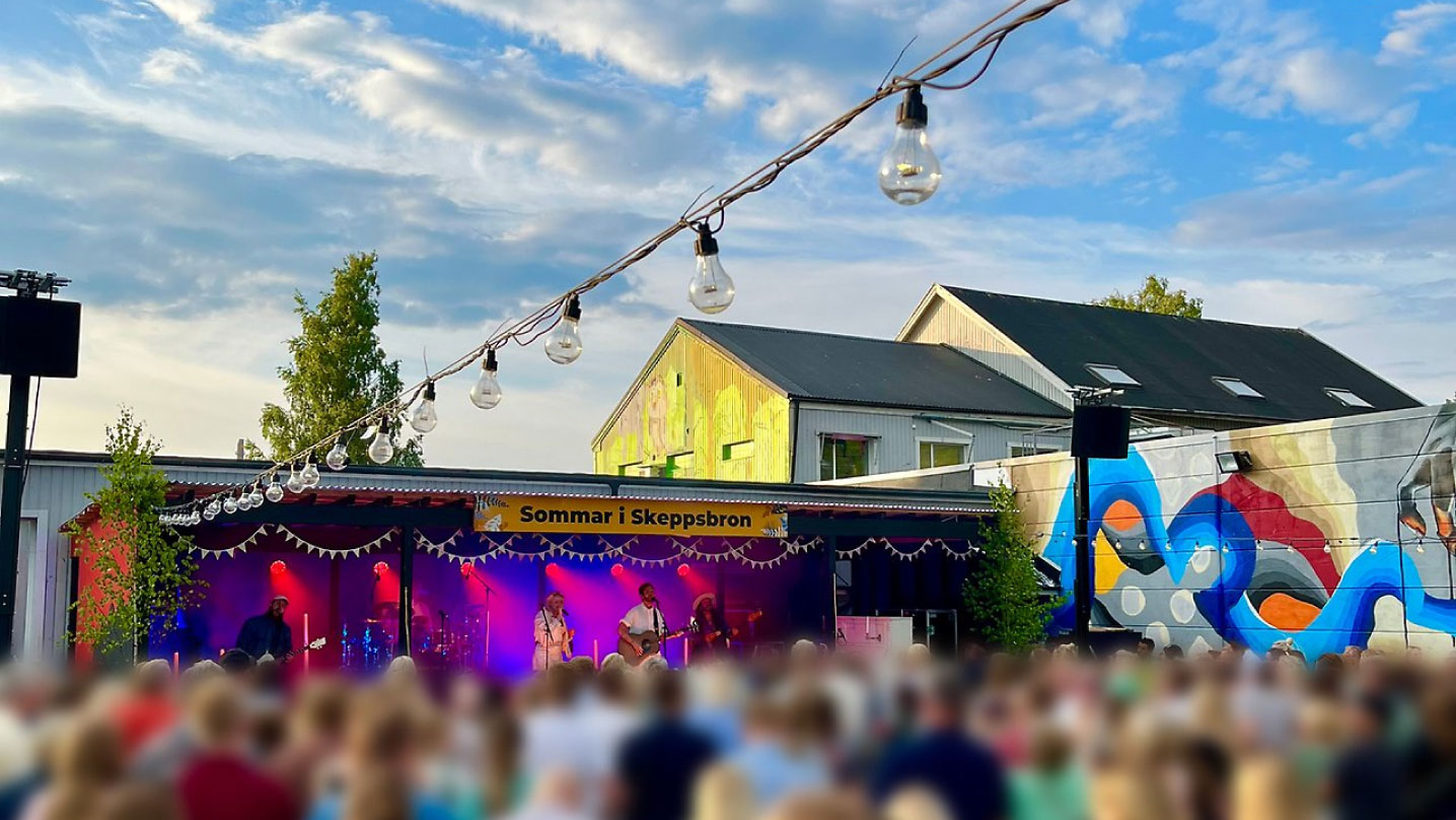 i förgrunden syns huvuden från en tät publik. publiken är vänd mot en scen med texten "sommar i skeppsbron" fäst i överkant. På scen spelar ett band. I luften hänger ljusslingor och runt om scenen finns färgglada väggar.