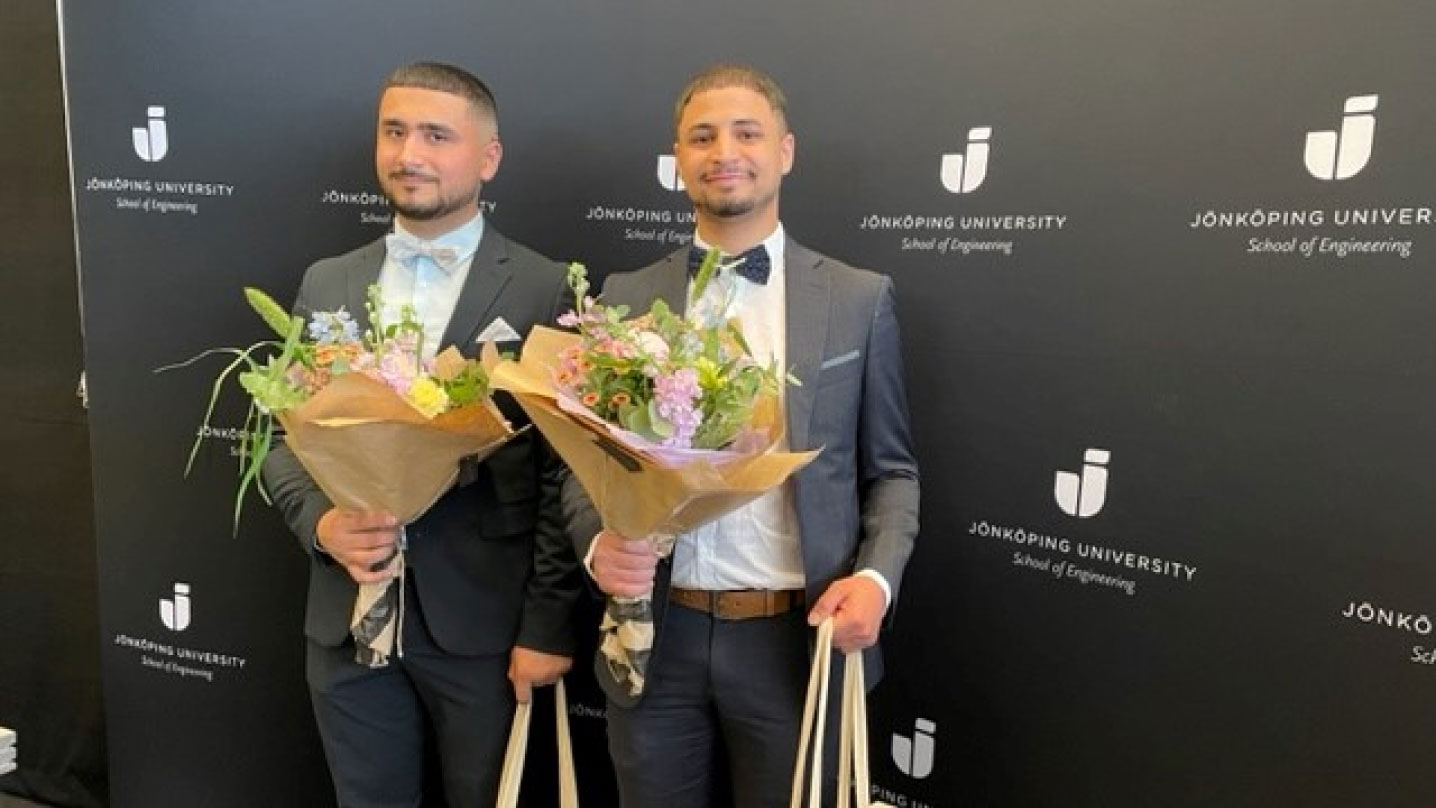 Två finklädda manliga stipendiater står framför en roll-up med Jönköping Universitets logga på. I handen håller de blommor och sina stipendier.