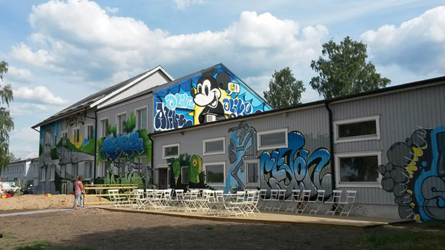 Ett stort grått trähus pyntat med några muralmålningar. Utanför huset står massa caféstolar.