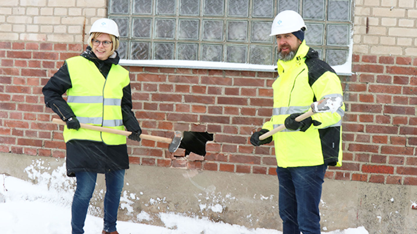 Två personer i varselväst står med hjälm och slägga framför ett tegelhus. De har slagit ett hål i väggen.
