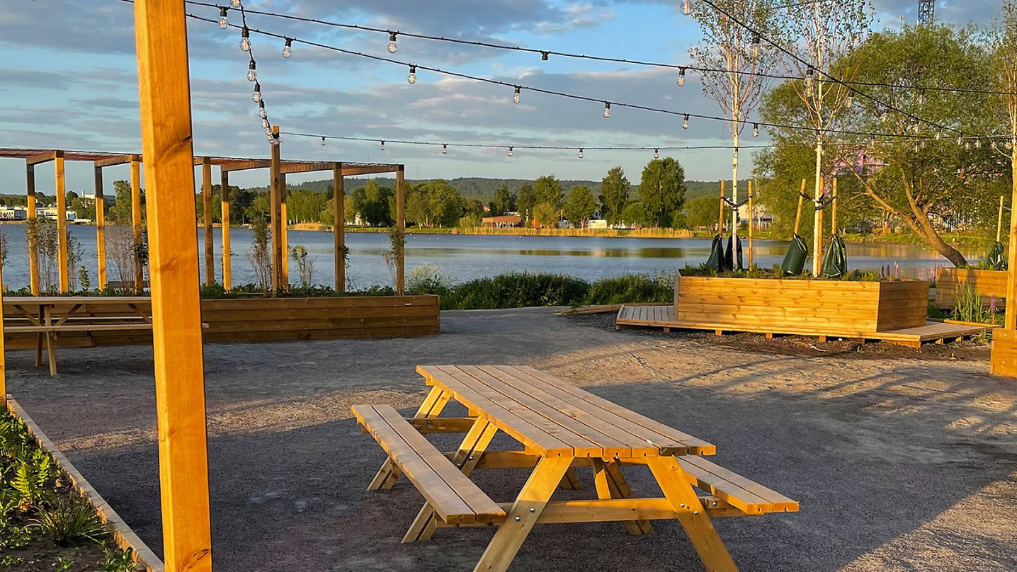 På en grusplan står ett campingbord i trä. Runt om finns flera blomlådor, sittplatser och pergolas i samma trä. Ljusslingor hänger i skyn och sjön ligger i bakgrunden.