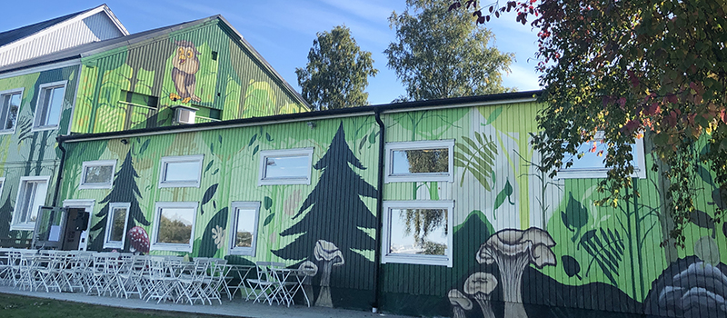 Foto på en del av byggnaden där Kvarteret Ödlan finns. Man ser en träfasad som är målad med en mängd figurer, djur, träd, svampar osv i gröna toner.