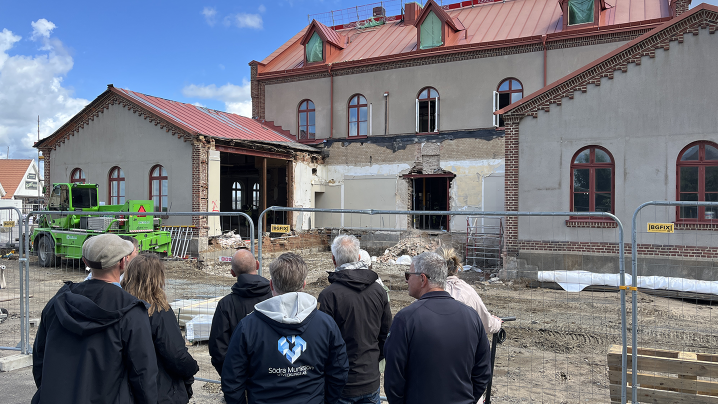 En grupp människor står utanför ett byggstängsel och tittar på en ljus byggnad som restaureras.