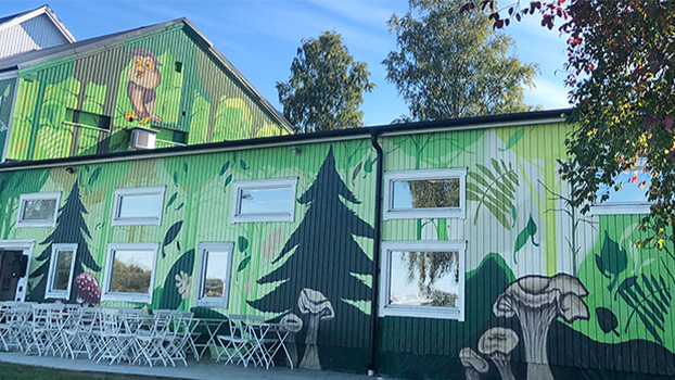 Ett hus är målat som en skog med granar och träd. Framför huset står vita stolar och cafébord.
