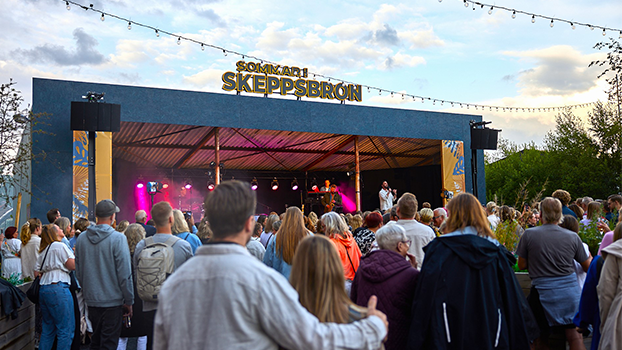 Ett folkhav står med ryggen mot kameran och tittar mot en stor scen. På scenens tak syns en stor gul ljusskylt med texten "Sommar i Skeppsbron".