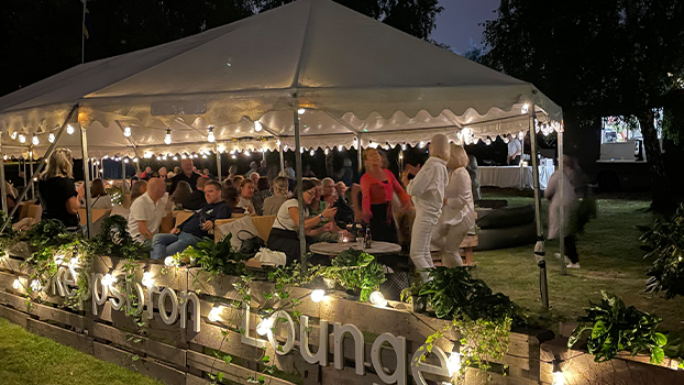 En lounge är uppbyggd under några stora träd. Loungen är inhägnad av ett staket av lastpallar och grönska och ljusslingor ligger ovanpå staketet. Inne i loungen sitter och står människor.
