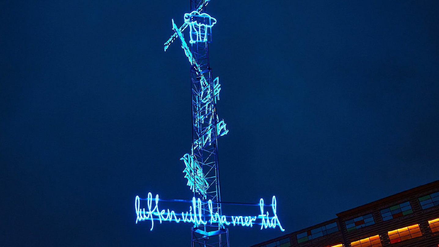 En mast som sträcker sig upp i skyn med flera olika neonslingor på. Slingorna är formade till olika föremål och texten "luften vill ha mer tid".
