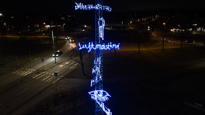 En mast sträcker sig i den mörka skyn, inklädd i flera neonslingor lysande i blått. Neonslingorna är formade i text och flera olika symboler.