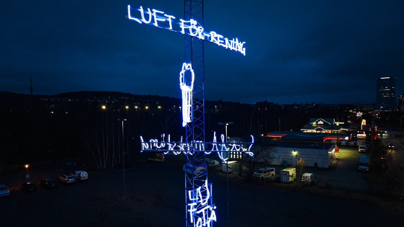 En mast sträcker sig i den mörka skyn, inklädd i flera neonslingor lysande i blått. Neonslingorna är formade i text och flera olika symboler.