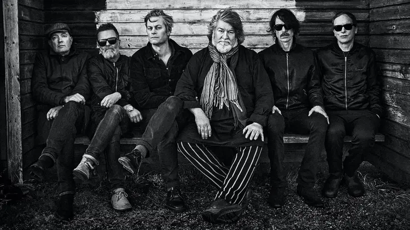 6 män sitter i mörka kläder på en bänk och tittar in i kameran. Bilden är svartvit.