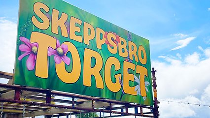 Fotografi av en färgglad skylt med grönt gräs och rosa, gula, röda blommor. Det står "Skeppsbrotorget" i gula bokstäver. Skylten sitter uppe på en byggställning och himlen är lysande blå i bakgrunden.