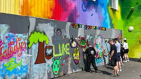 Flera personer testar på att måla graffiti på en vägg. Väggen är fylld med färgglada motiv och texter.
