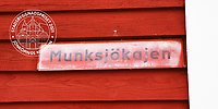 En gammal gatuskylt med text "Munksjökajen" på en faluröd trävägg.