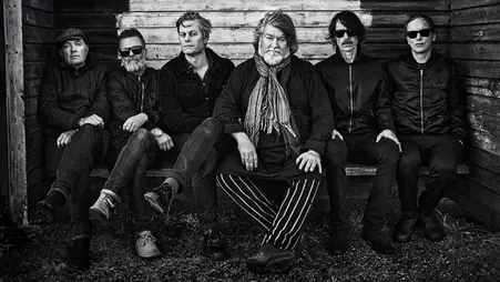 6 män sitter i mörka kläder på en bänk och tittar in i kameran. Bilden är svartvit.