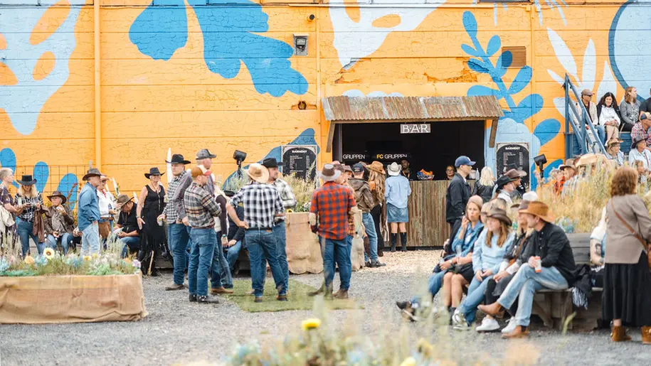 Människor i cowboyhatt och rutig skjorta står utspridda framför en orange vägg.