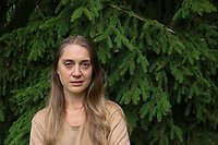 Konstnären Hanna Wildow står framför en grön barrskog. Hon har beige t-shirt och långt ljusbrunt hår.